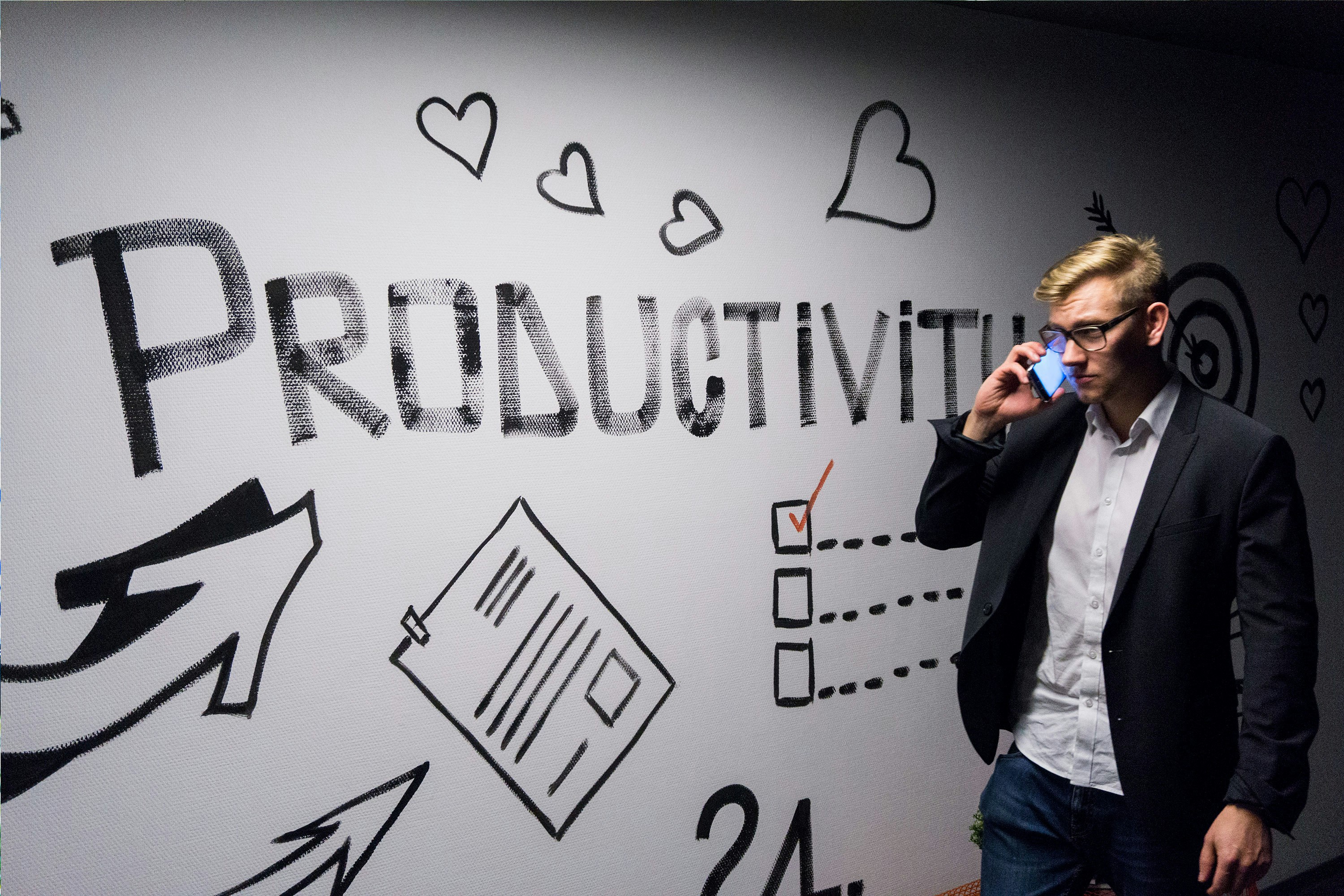 En mann i dressjakke snakker i telefonen foran en vegg med teksten «PRODUCTIVITY» og håndtegnede ikoner.